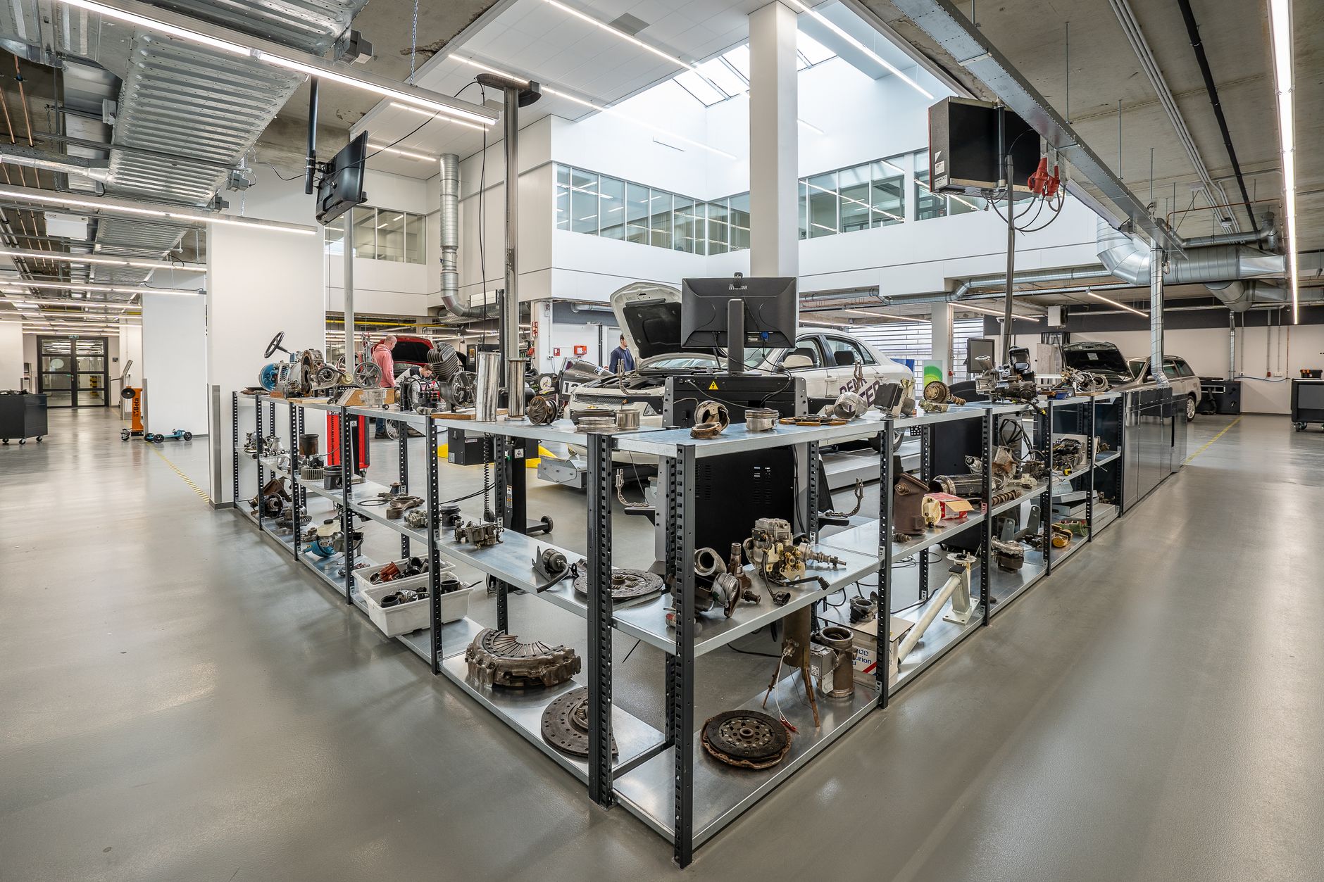 Atelier industriel avec étagères métalliques chargées de pièces mécaniques, écrans d’ordinateur et voitures en arrière-plan.