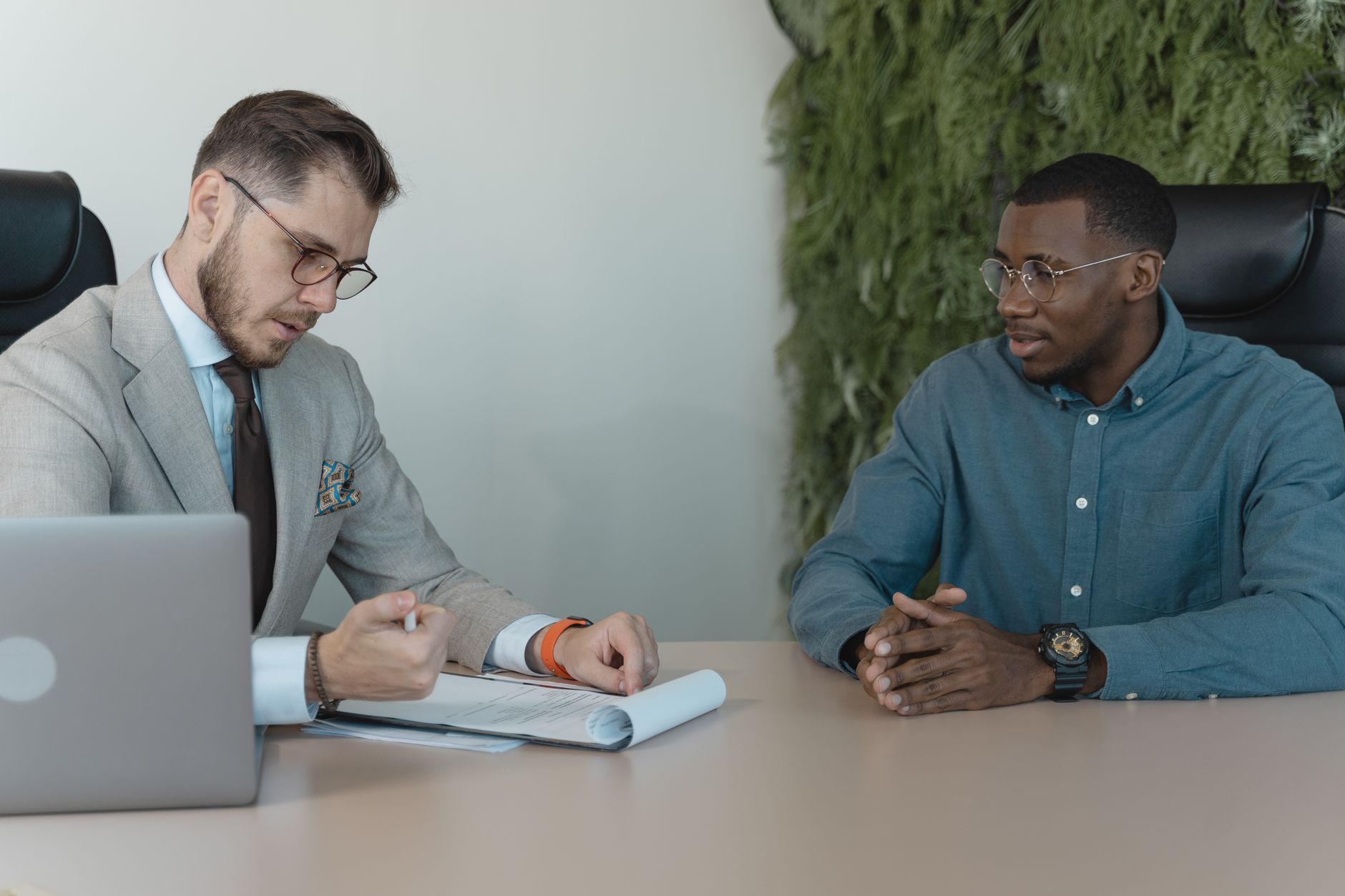 Deux hommes à une table de bureau : l’un consulte des documents, l’autre écoute les mains jointes, devant un mur végétal.