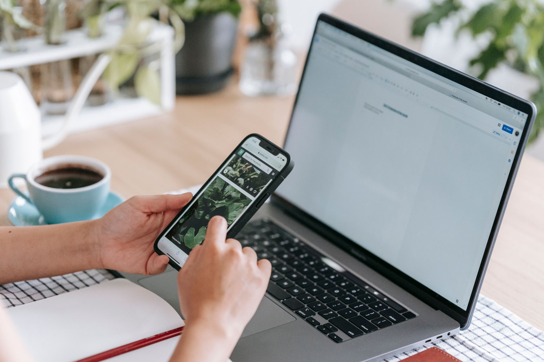 Deux mains tiennent un smartphone devant un ordinateur portable ouvert, sur un bureau avec une tasse de café, un carnet et des plantes.