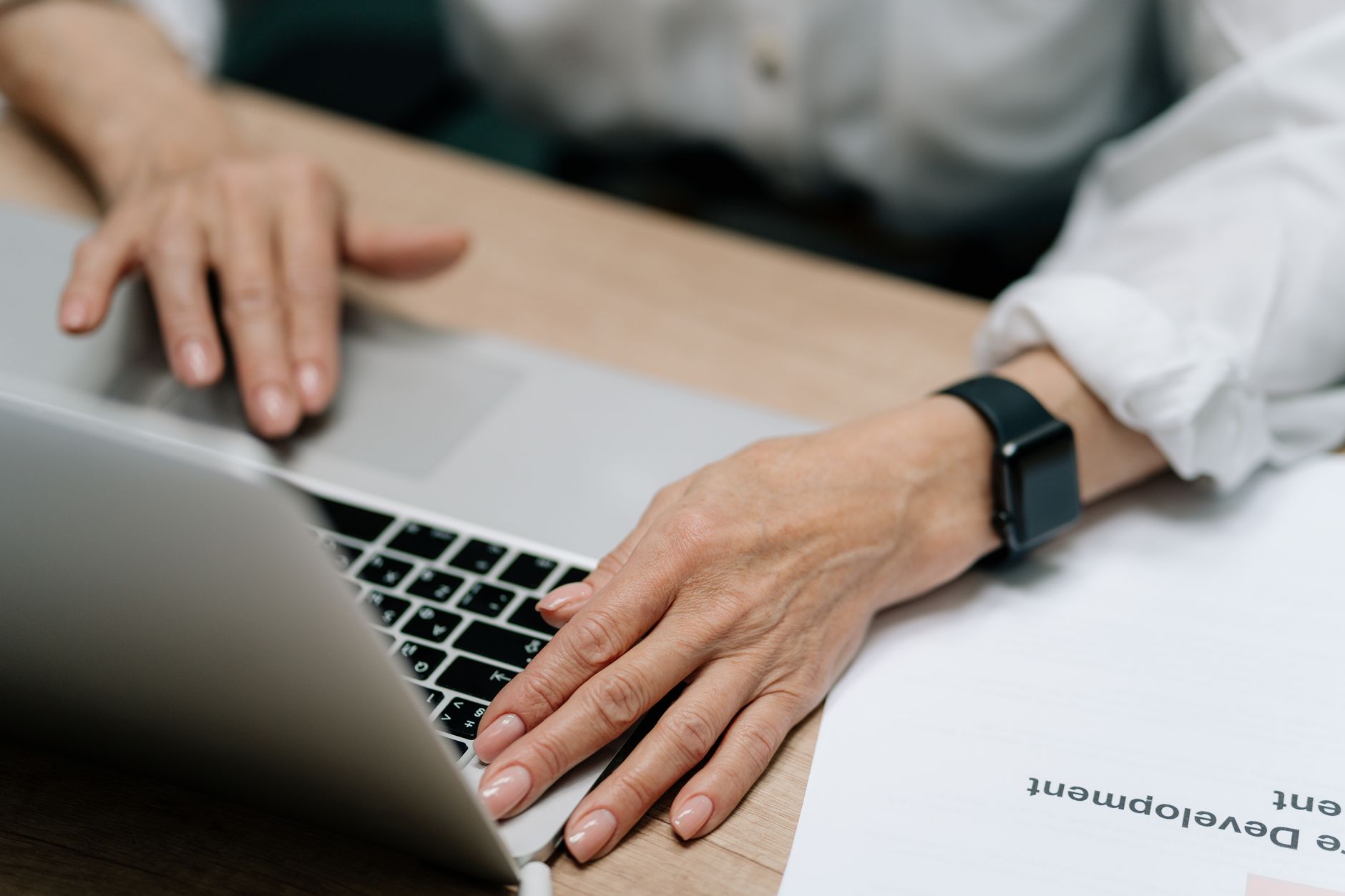 Mains d’une personne en chemise blanche utilisant un ordinateur portable sur un bureau en bois, avec une montre noire au poignet et une feuille à côté.
