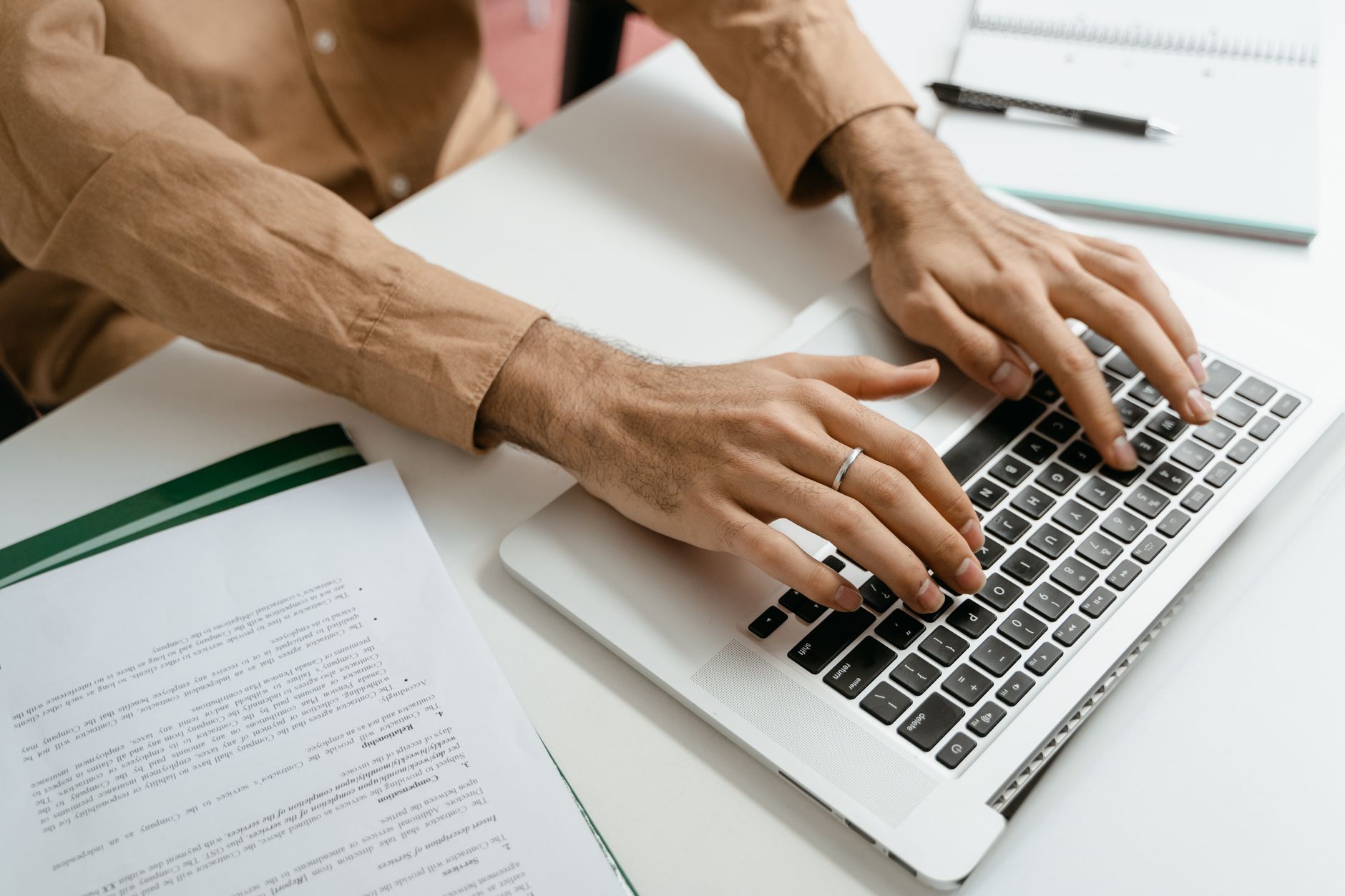 Mains d’une personne tapant sur un ordinateur portable, sur un bureau blanc avec des documents imprimés, un carnet et un stylo.