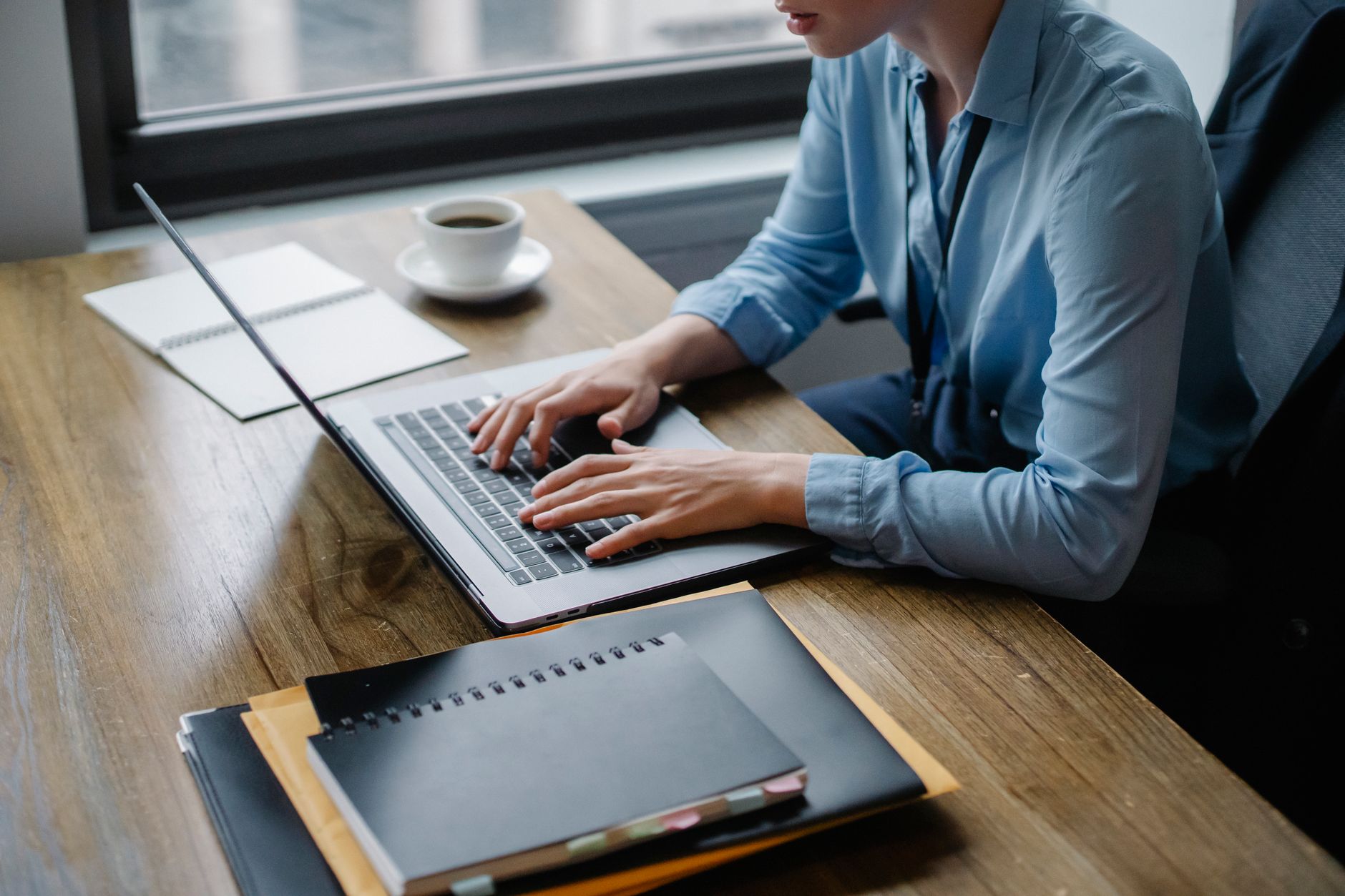 Personne en chemise bleue tapant sur un ordinateur portable à une table en bois, avec une tasse de café, des carnets et des dossiers.